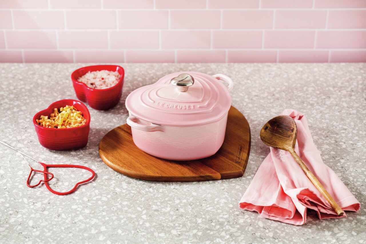 LE CREUSET Herzbräter pink 20 cm HEART COLLECTION Herzförmiger, pinker Bräter von Le Creuset auf einem herzförmigen Holzbrett, umgeben von roten herzförmigen Schalen mit Zutaten, aus der Vogelperspektive fotografiert.