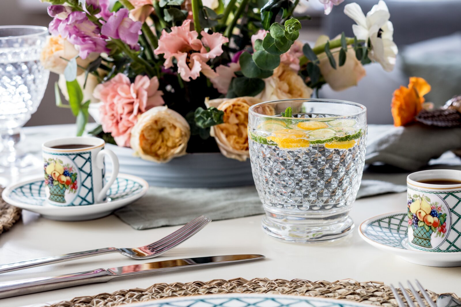 Een stijlvol gedekte tafel met op de voorgrond een Boston-beker, gevuld met water, schijfjes citroen en limoen, omringd door bloemen en serviesgoed. Vooraanzicht.