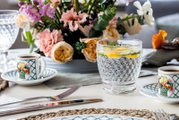 Een stijlvol gedekte tafel met op de voorgrond een Boston-beker, gevuld met water, schijfjes citroen en limoen, omringd door bloemen en serviesgoed. Vooraanzicht.