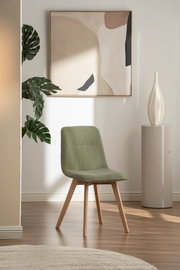Moderner grüner Stuhl mit Holzbeinen, frontal fotografiert in einem stilvoll eingerichteten Raum mit Kunstwerk und Pflanze im Hintergrund.