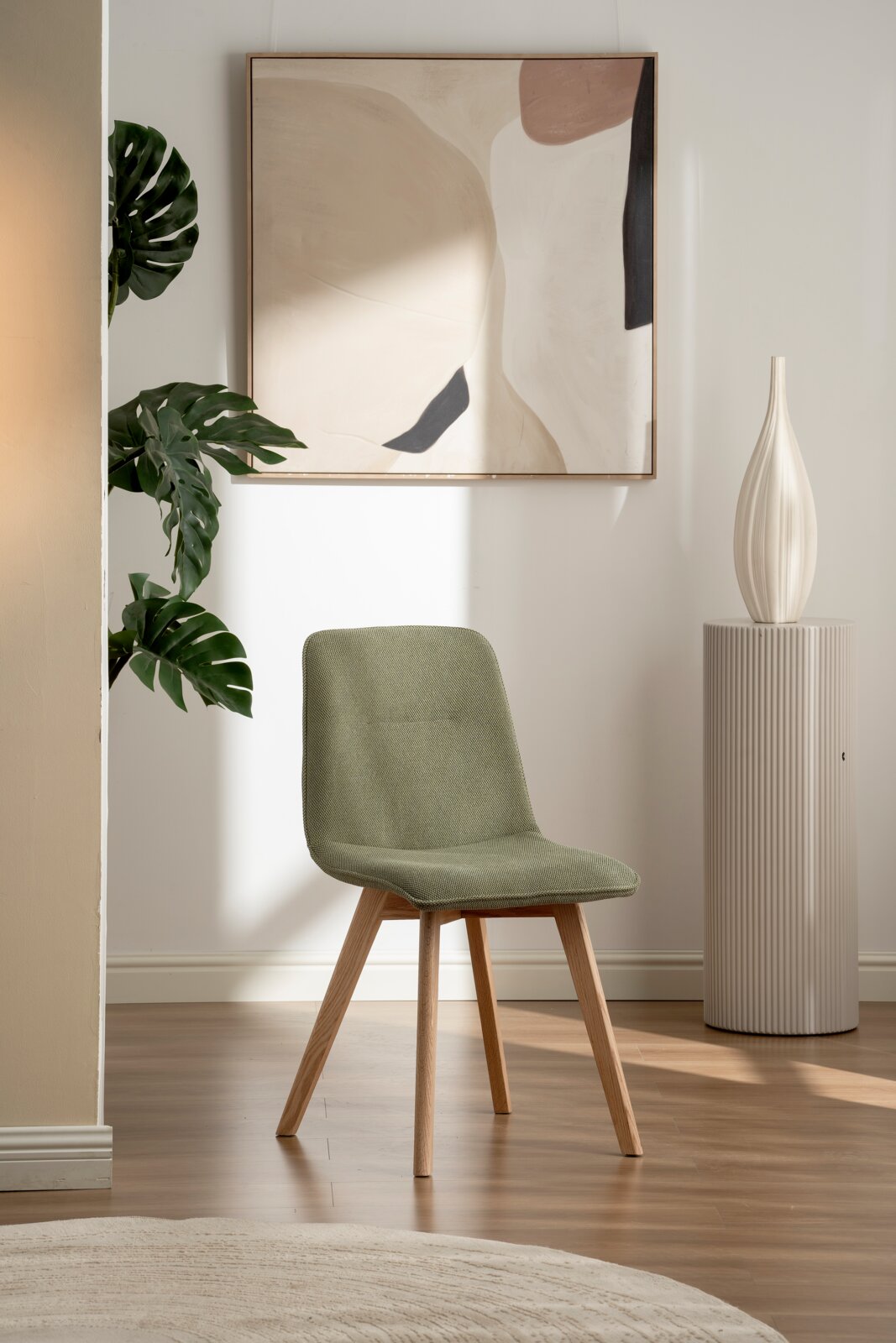 Moderner grüner Stuhl mit Holzbeinen, frontal fotografiert in einem stilvoll eingerichteten Raum mit Kunstwerk und Pflanze im Hintergrund.