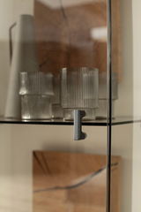 Nahaufnahme einer Glastür mit Metallgriff, dahinter befinden sich geriffelte Gläser und eine Vase. Perspektive von vorne.