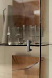 Nahaufnahme einer Glastür mit Metallgriff, dahinter befinden sich geriffelte Gläser und eine Vase. Perspektive von vorne.