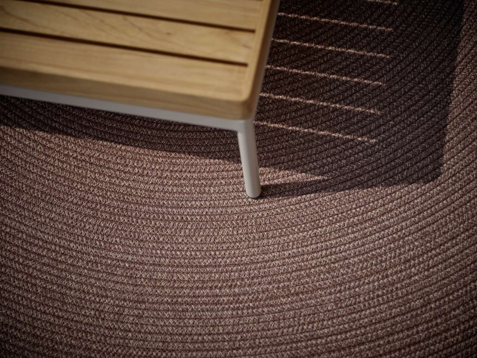 Nahaufnahme eines Outdoor-Teppichs mit geflochtener Struktur in Braun, teilweise von einem Holztisch beschattet.