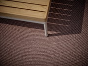 Close-up van een buitentapijt met gevlochten structuur in bruin, gedeeltelijk in de schaduw van een houten tafel.