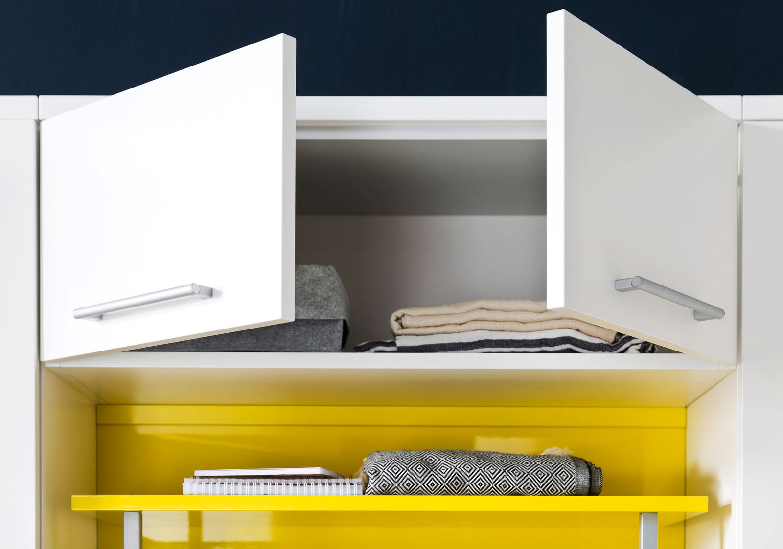 Offener weißer Schrank mit zwei Türen, gefüllt mit gefalteten Handtüchern und einem Notizbuch auf einem gelben Regal. Frontalansicht.