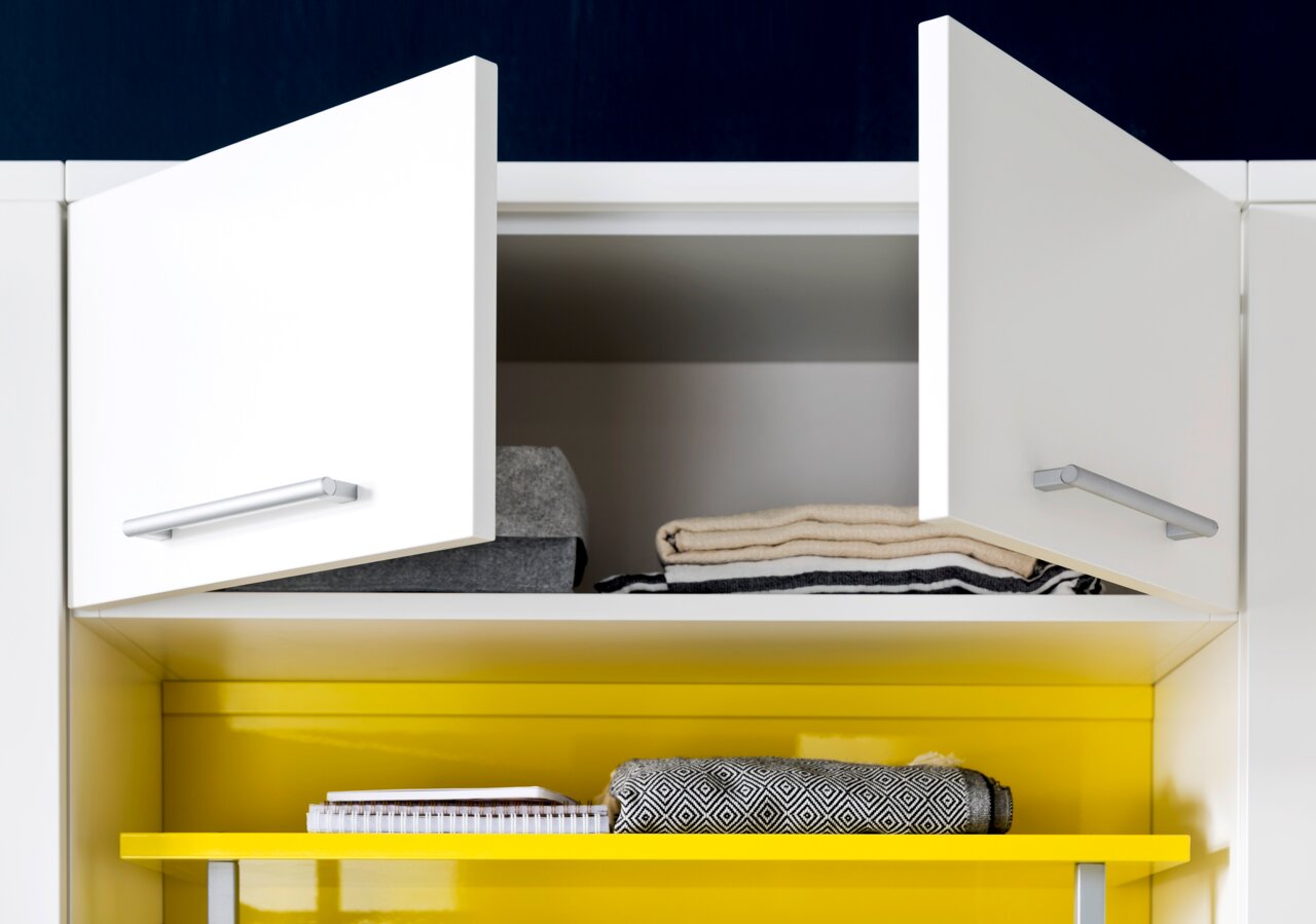 Offener weißer Schrank mit zwei Türen, gefüllt mit gefalteten Handtüchern und einem Notizbuch auf einem gelben Regal. Frontalansicht.