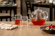 Glas-Teekanne mit integriertem Sieb und zwei Glastassen, gefüllt mit rotem Tee, auf einem Holztisch. Perspektive: seitlich.