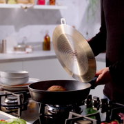Iemand tilt het deksel van een Adamant Comfort-pan van 24 cm op een gasfornuis in een moderne keuken, gezien vanuit de zijkant.