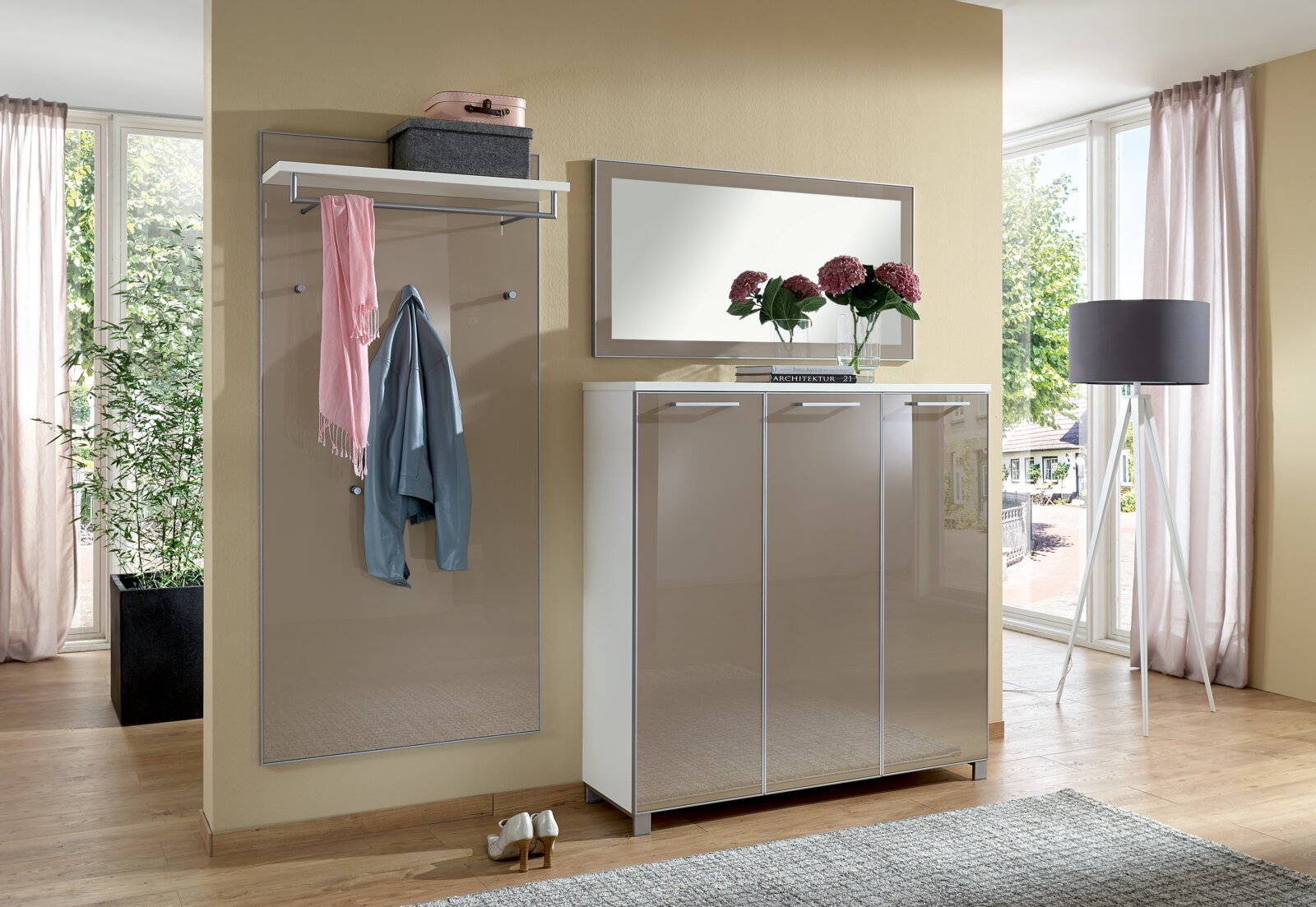 Modernes Garderoben-Set in einem hellen Raum mit großem Fenster, bestehend aus einem Spiegel, einer Garderobenleiste mit Ablage und einem dreitürigen Schrank. Perspektive von vorne.