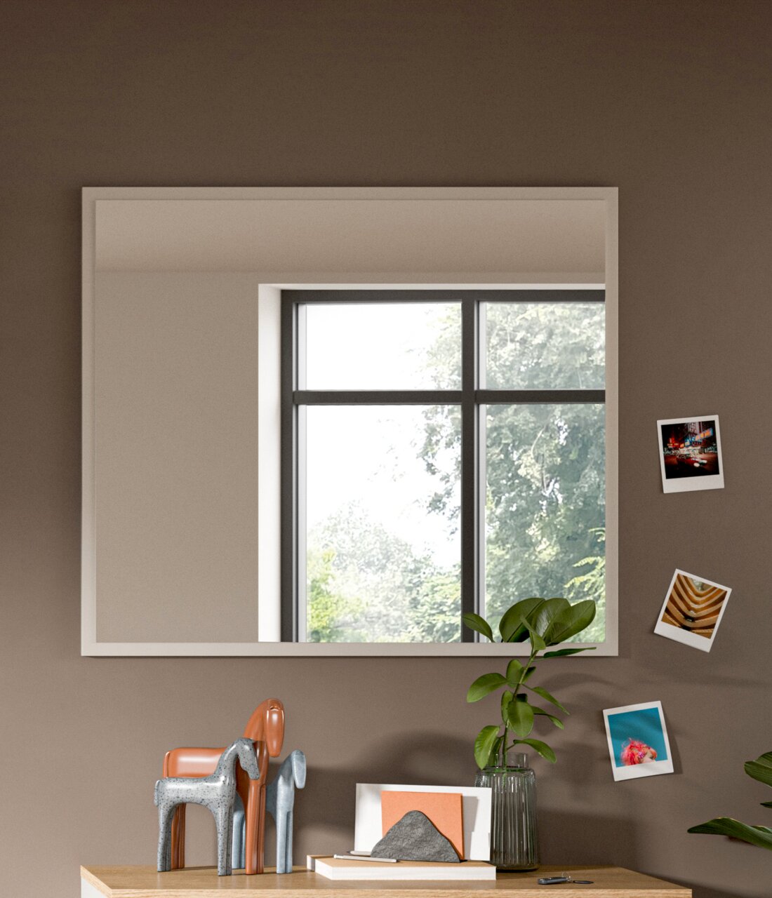 Moderner rechteckiger Spiegel an einer Wand, reflektiert ein Fenster mit Blick auf Bäume, dekoriert mit kleinen Fotos und Pflanzen, Perspektive von vorne.