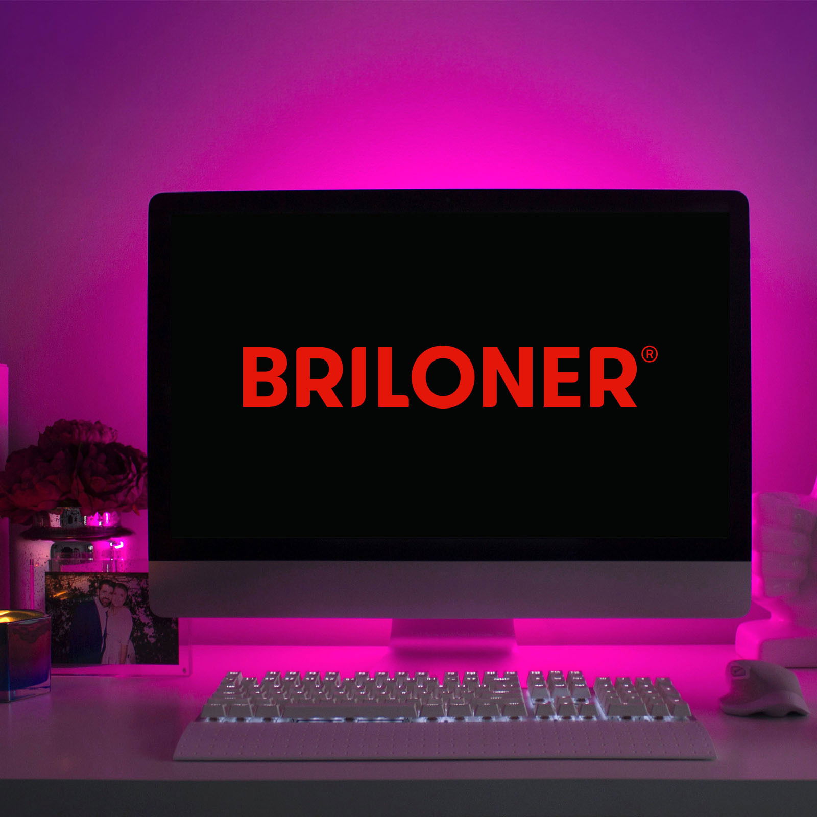 Briloner USB RGB-LED Flexband 4m Indoor Ein Computerbildschirm mit dem Logo 'BRILONER' in roter Schrift, umgeben von einem pinken LED-Licht, das eine stimmungsvolle Beleuchtung erzeugt. Perspektive: Frontalansicht auf den Bildschirm und die beleuchtete Umgebung.