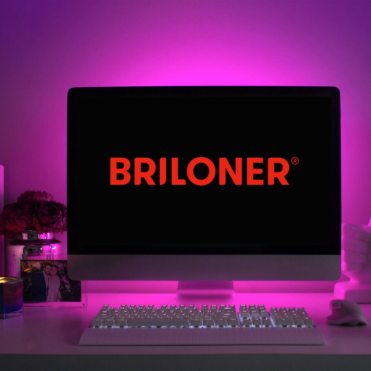 Ein Computerbildschirm mit dem Logo 'BRILONER' in roter Schrift, umgeben von einem pinken LED-Licht, das eine stimmungsvolle Beleuchtung erzeugt. Perspektive: Frontalansicht auf den Bildschirm und die beleuchtete Umgebung.