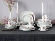 Frontale Tischansicht eines weißen Porzellan-Geschirrsets mit rosa Orchideenmotiv, bestehend aus Tellern, Schalen und Tassen, dekoriert mit Kerzenständern und Blumen auf grauem Hintergrund.