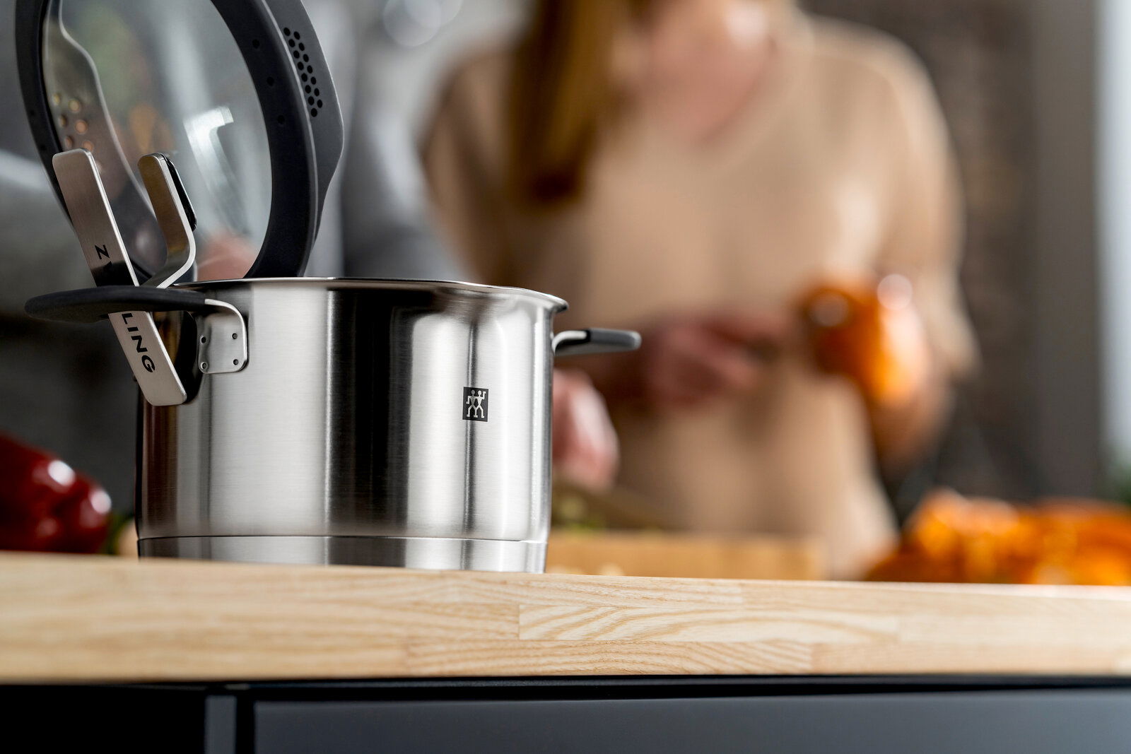 Nahaufnahme eines Edelstahltopfs mit Glasdeckel auf einer Küchenarbeitsplatte, im Hintergrund unscharf eine Person beim Kochen.
