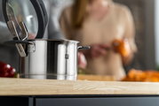 Nahaufnahme eines Edelstahltopfs mit Glasdeckel auf einer Küchenarbeitsplatte, im Hintergrund unscharf eine Person beim Kochen.