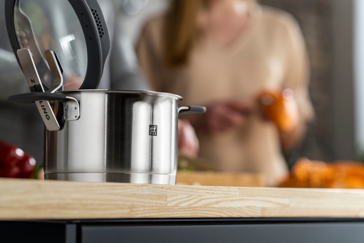 Close-up van een roestvrijstalen pan met glazen deksel op een keukenblad, op de achtergrond een onscherp beeld van iemand die aan het koken is.