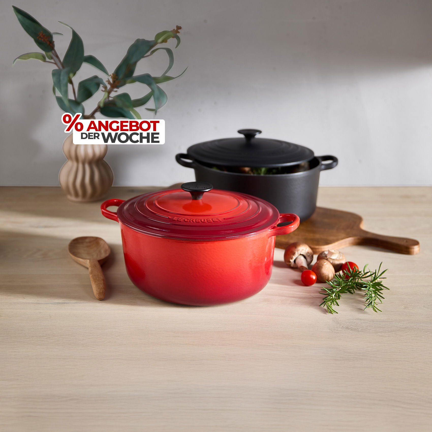 Roter und schwarzer Schmortopf auf Holztisch mit Kräutern und Gemüse, daneben Schriftzug „% Angebot der Woche“