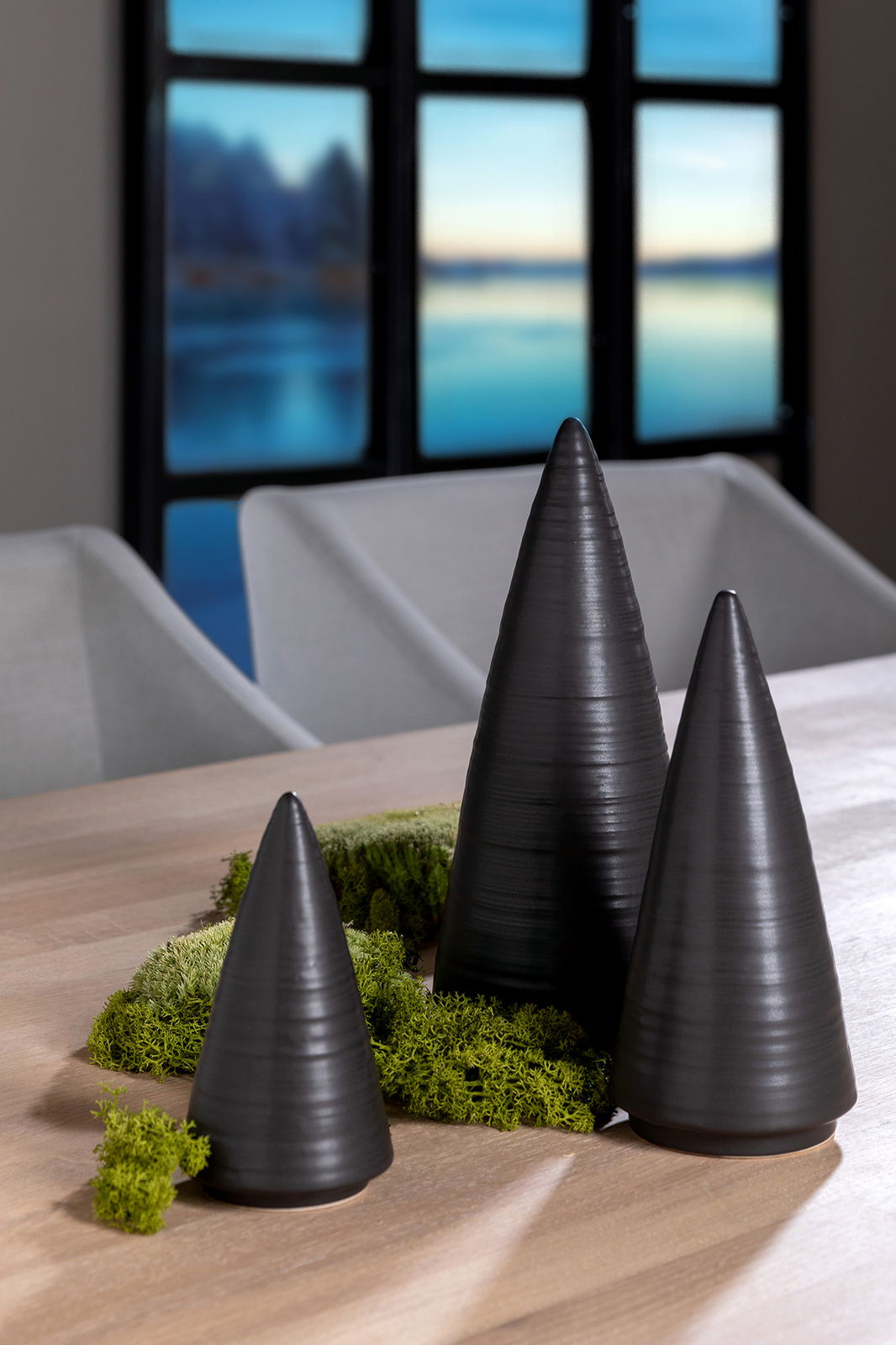 Drei schwarze, konische Dekobäume aus Keramik auf einem Holztisch, umgeben von grünem Moos, mit einem Fenster im Hintergrund. Perspektive: seitlich.