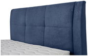 Nahaufnahme eines blauen Boxspringbett-Kopfteil mit gepolstertem Design und einer weißen Matratze im Vordergrund, seitliche Perspektive.