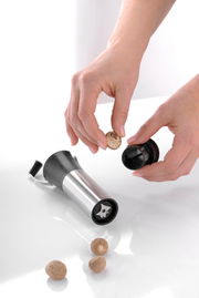 Muskatmühle MOSCATA LASER CUT aus Edelstahl, liegend auf einem Tisch, mit einer Hand, die eine Muskatnuss in die Mühle einlegt. Perspektive von oben.