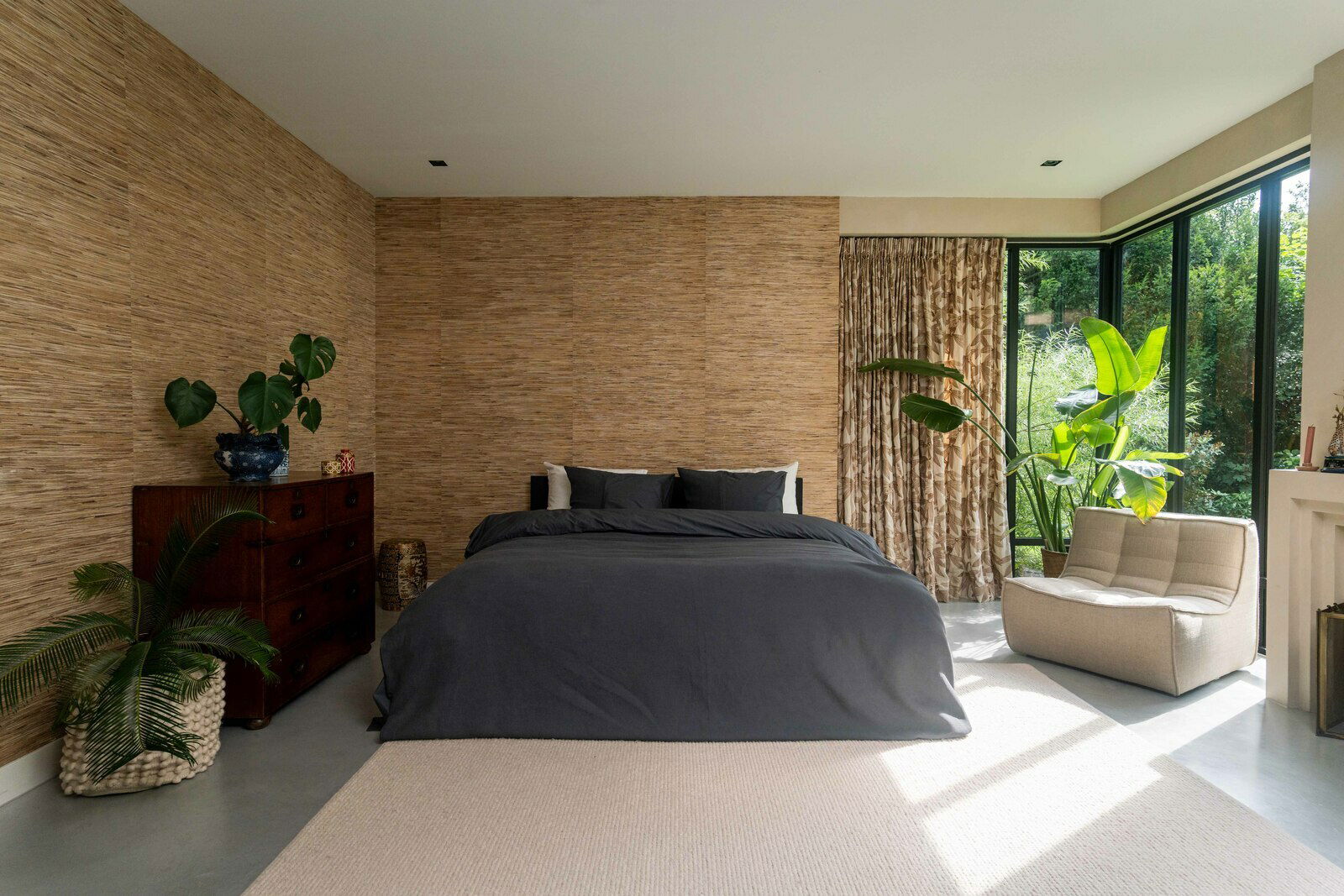 Modernes Schlafzimmer mit einem großen Bett in dunkelgrauer Bettwäsche, umgeben von Pflanzen und natürlichem Licht von großen Fenstern, aus der Perspektive von vorne.