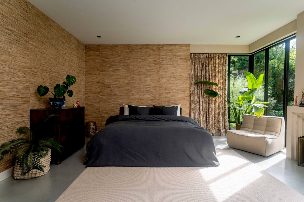 Modernes Schlafzimmer mit einem großen Bett in dunkelgrauer Bettwäsche, umgeben von Pflanzen und natürlichem Licht von großen Fenstern, aus der Perspektive von vorne.