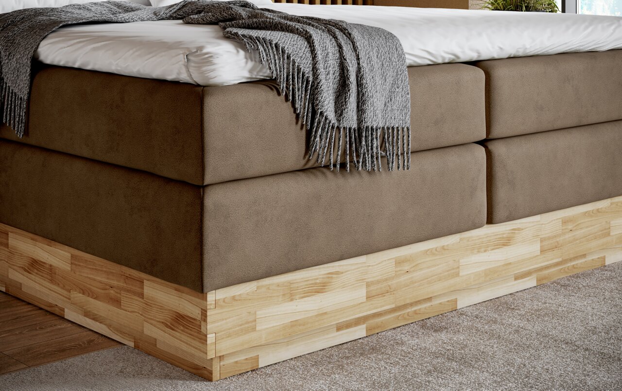 Close-up van een boxspringbed met bruine stoffen bekleding en houten onderstel, gezien vanaf de zijkant.