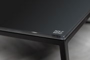 Nahaufnahme eines eleganten Couchtischs mit schwarzer Glasplatte und Metallrahmen, Markenlogo in der Ecke sichtbar.
