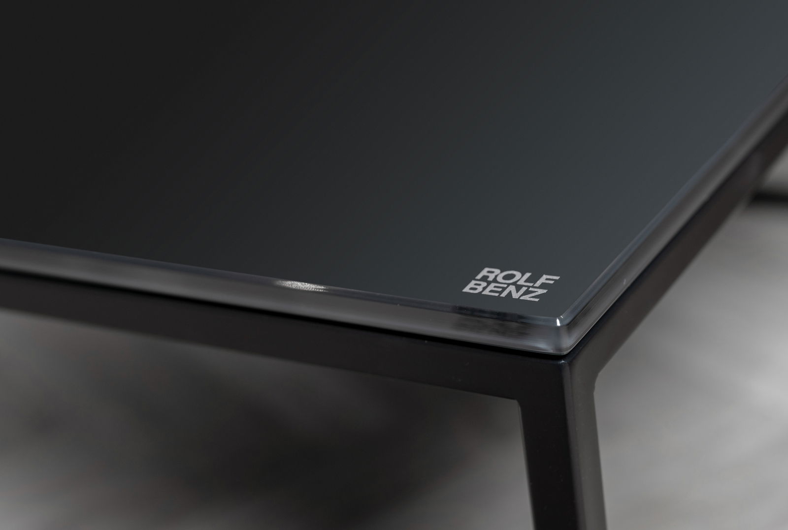 Nahaufnahme eines eleganten Couchtischs mit schwarzer Glasplatte und Metallrahmen, Markenlogo in der Ecke sichtbar.
