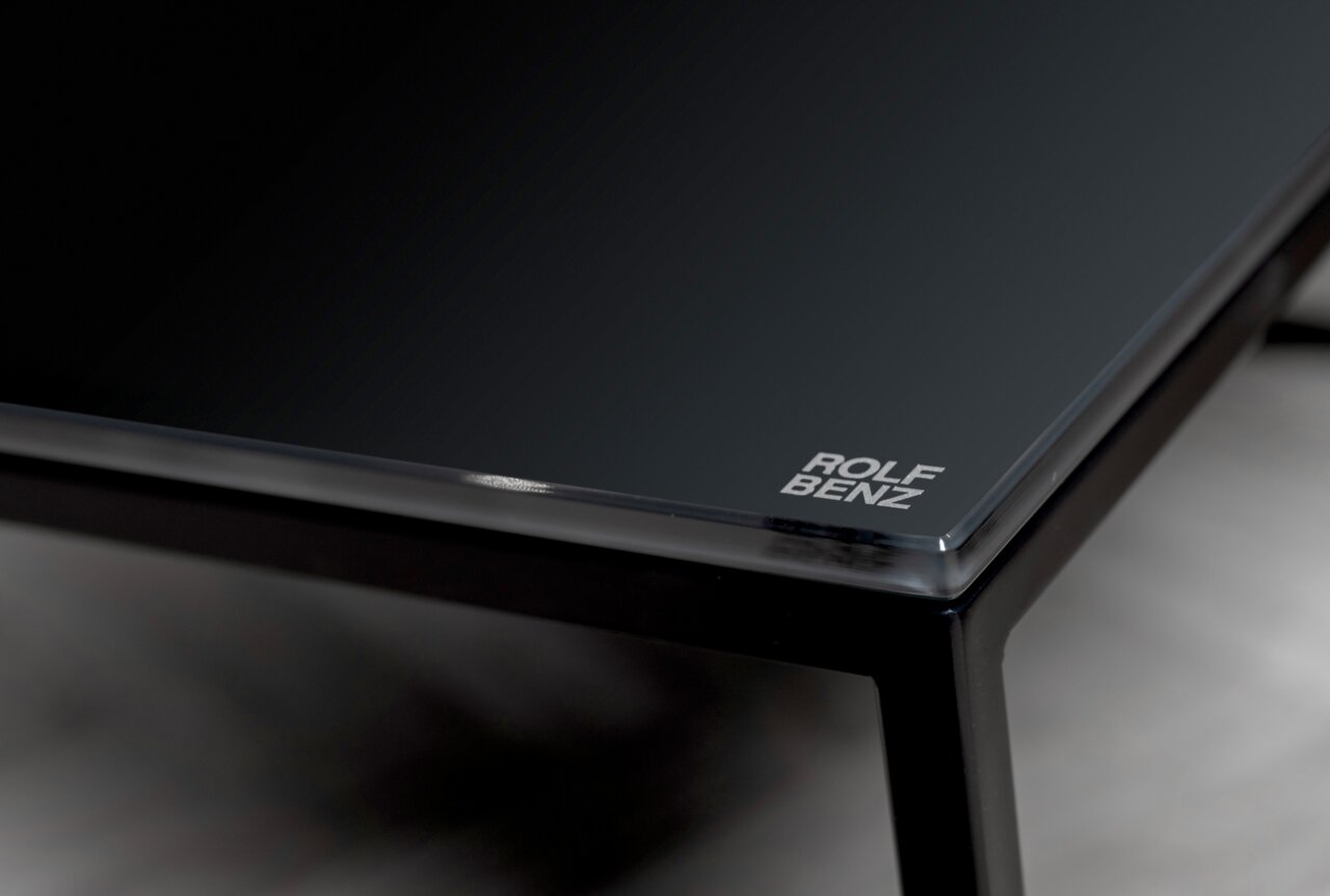 Nahaufnahme eines eleganten Couchtischs mit schwarzer Glasplatte und Metallrahmen, Markenlogo in der Ecke sichtbar.