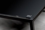 Close-up van een elegante salontafel met een zwart glazen blad en een metalen frame; het merklogo is in de hoek te zien.