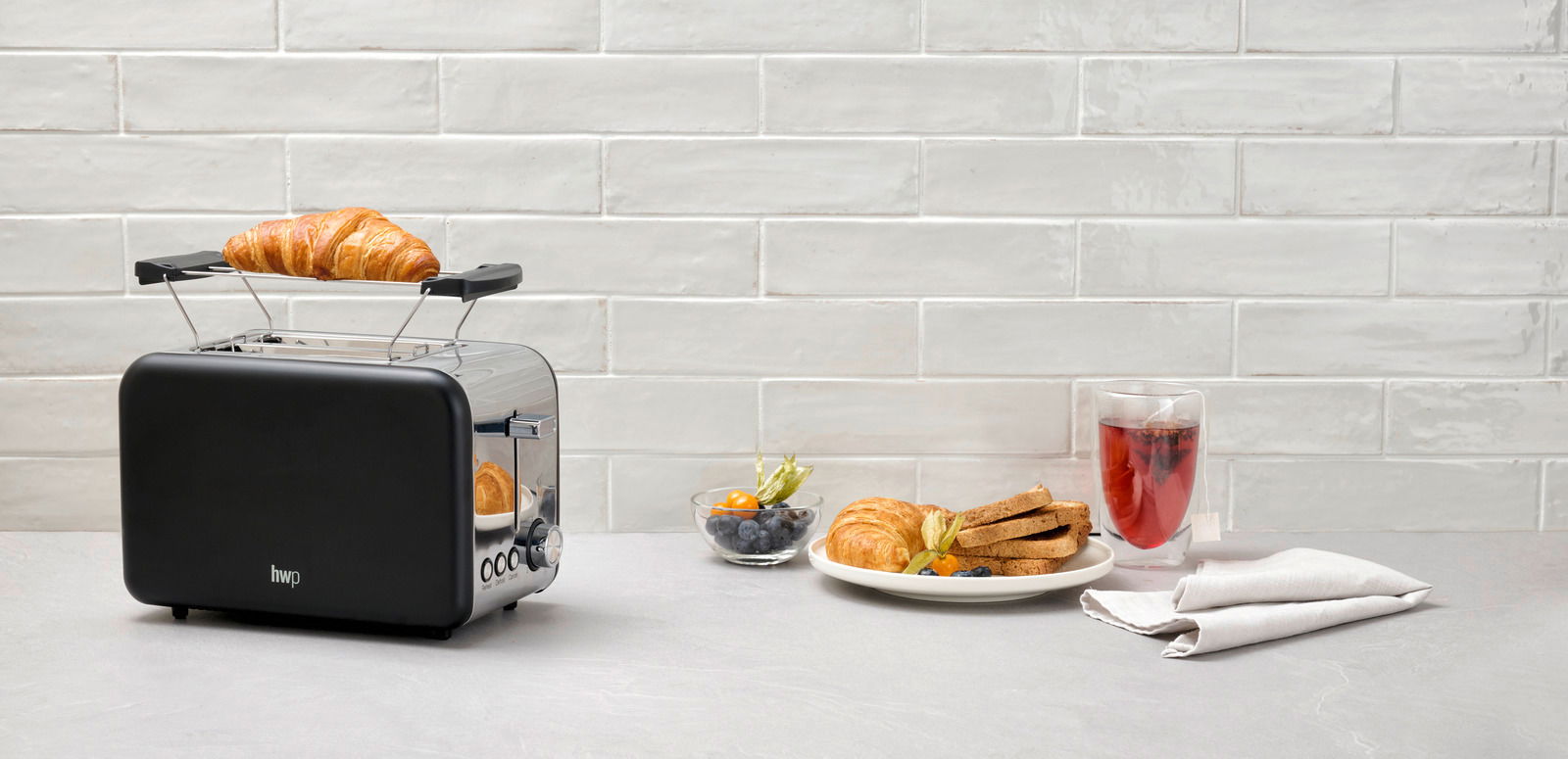 Schwarzer Toaster mit einem Croissant auf dem Brötchenaufsatz, seitliche Perspektive, daneben ein Teller mit Toast und Beeren.