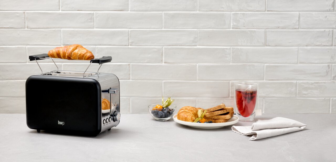 Schwarzer Toaster mit einem Croissant auf dem Brötchenaufsatz, seitliche Perspektive, daneben ein Teller mit Toast und Beeren.