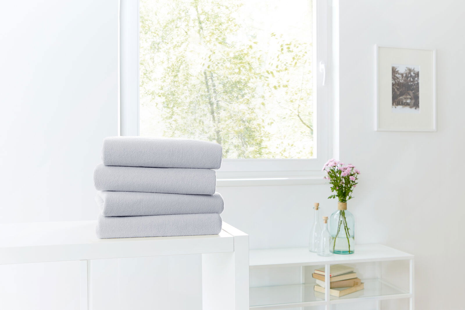 Ein Stapel grauer Spannbetttücher auf einem weißen Tisch, im Hintergrund ein Fenster mit Blick ins Grüne, seitliche Perspektive.