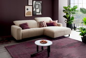 Beiges Ecksofa mit rechter Ottomane und schwarzen Metallfüßen in modernem Wohnzimmer, fotografiert in leichter Schrägansicht von vorn; mit Zierkissen, Couchtisch und Pflanzen am Fenster.