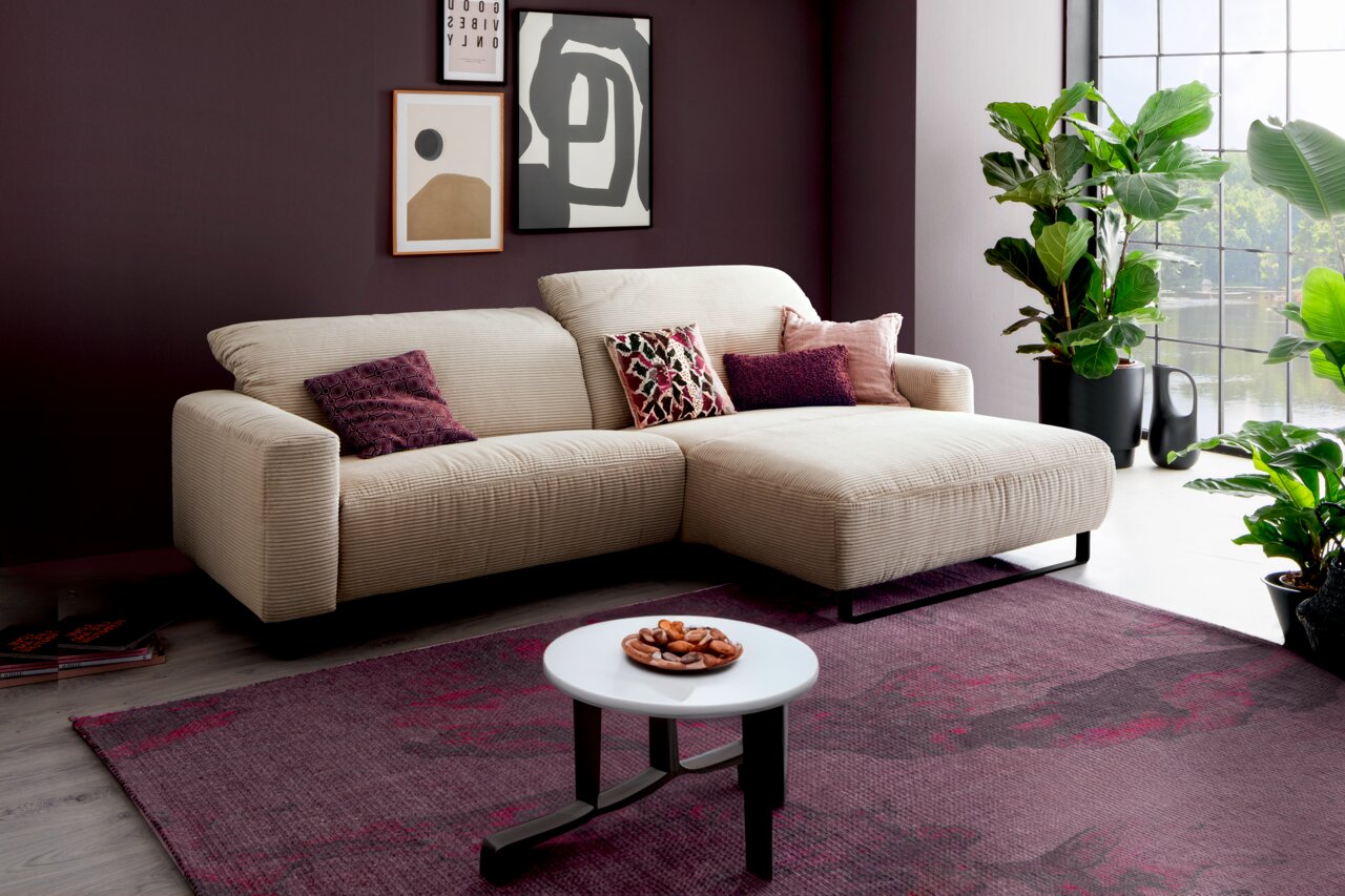 Beiges Ecksofa mit rechter Ottomane und schwarzen Metallfüßen in modernem Wohnzimmer, fotografiert in leichter Schrägansicht von vorn; mit Zierkissen, Couchtisch und Pflanzen am Fenster.