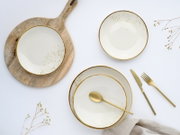 Draufsicht auf ein elegantes Geschirr-Set aus weißen Schalen und Tellern mit goldfarbenem Rand und Sprenkelmuster, arrangiert auf weißem Hintergrund mit Holzbrett sowie goldenem Besteck.