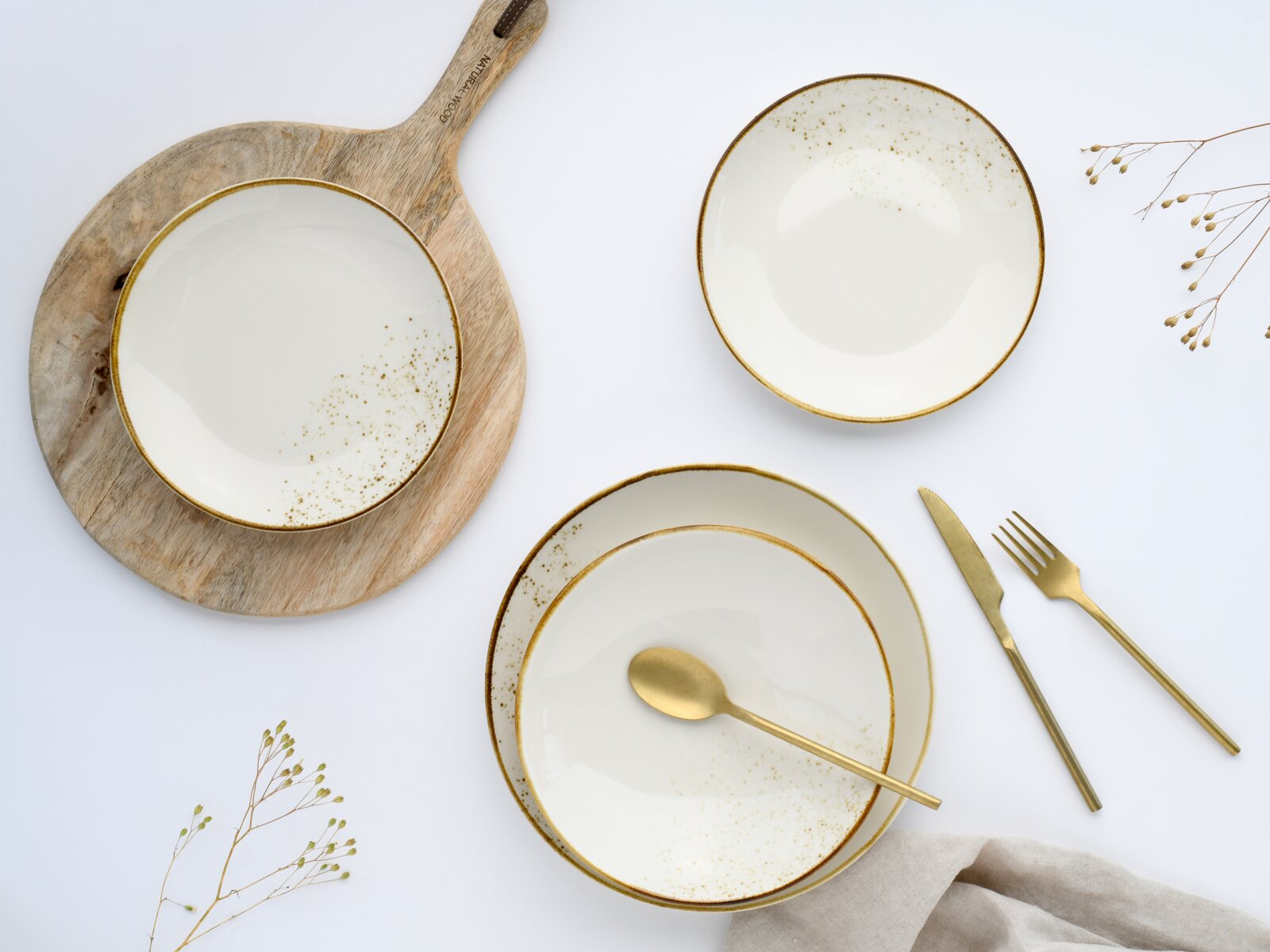 Draufsicht auf ein elegantes Geschirr-Set aus weißen Schalen und Tellern mit goldfarbenem Rand und Sprenkelmuster, arrangiert auf weißem Hintergrund mit Holzbrett sowie goldenem Besteck.