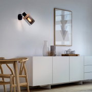 Schwarze Wandlampe mit zylindrischem Rauchglas-Schirm und Holzdetail, eingeschaltet, seitliche Ansicht an heller Wand in modernem Wohnraum