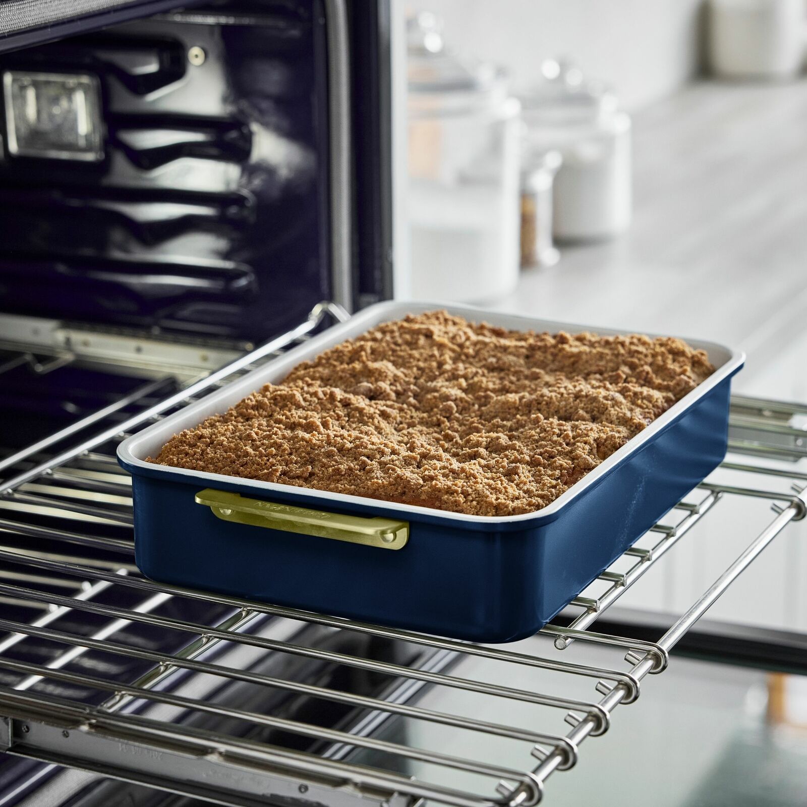 Rechteckige Backform mit blauem Rand und goldenen Griffen, gefüllt mit einem gebackenen Streuselkuchen, auf einem Ofengitter von der Seite fotografiert.