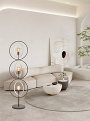 Moderne bruine vloerlamp met drie verlichte glazen ringen, vanuit schuin vooraanzicht in een lichte, minimalistische woonkamer