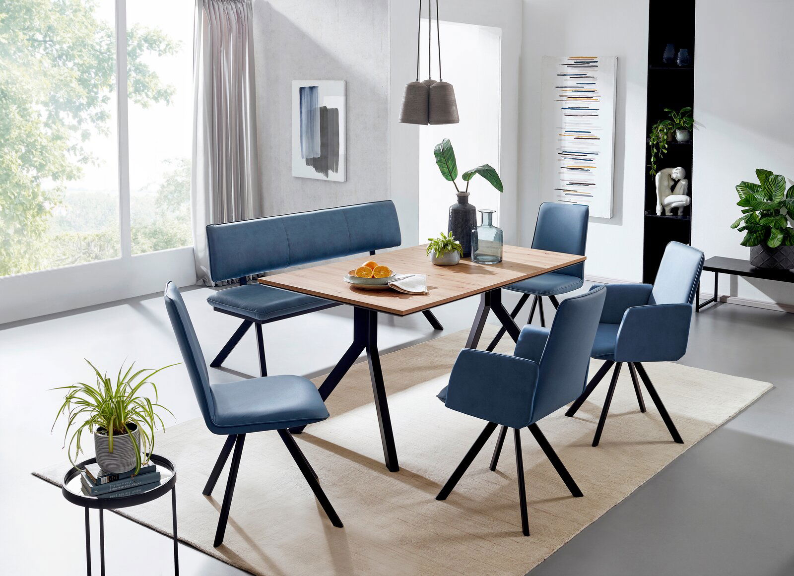 Moderner Vierfußtisch aus Holz mit schwarzem Metallgestell, umgeben von blauen Stühlen und einer Bank, in einem hellen Esszimmer mit Pflanzen und Dekorationen. Perspektive: seitlich von oben.