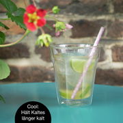 Ein doppelwandiges Glas mit einem Getränk, Eiswürfeln, Limettenscheiben und einem Strohhalm, auf einem blauen Tisch vor einer Ziegelwand. Perspektive: seitlich.