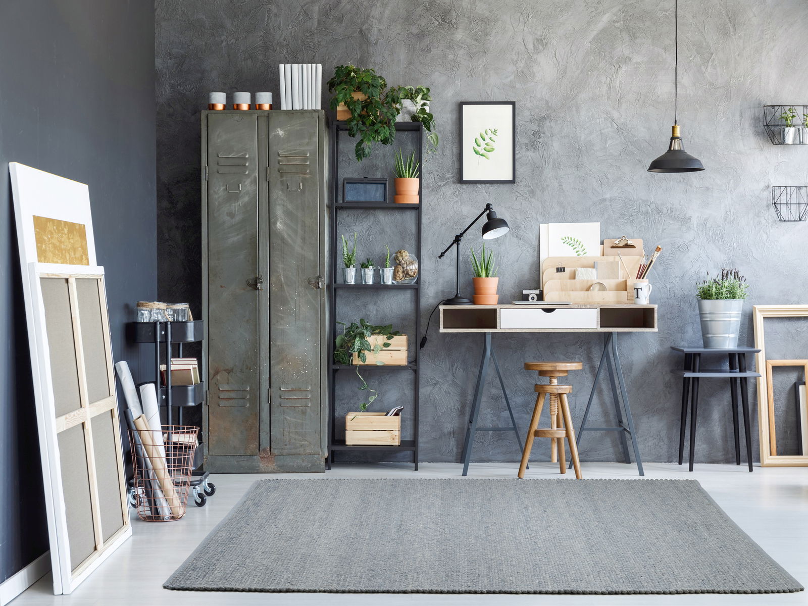 Moderner Raum mit grauem Handweb Teppich im Vordergrund, umgeben von industriellen und natürlichen Elementen wie einem Metallschrank, Pflanzen und einem Schreibtisch mit Dekorationen.
