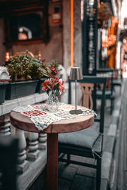 Akku-Tischleuchte VILETTO auf einem runden Holztisch mit gehäkeltem Tischläufer und Blumenvase, seitliche Perspektive.
