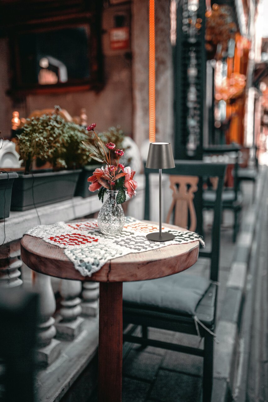 Akku-Tischleuchte VILETTO auf einem runden Holztisch mit gehäkeltem Tischläufer und Blumenvase, seitliche Perspektive.