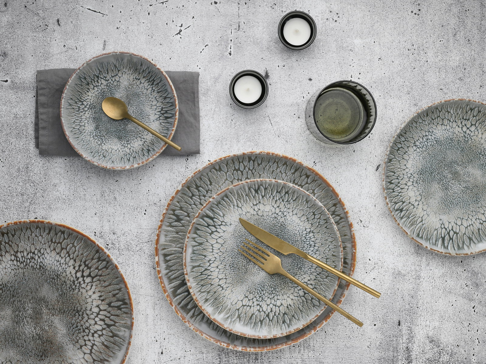 Vogelperspektive auf ein 12-teiliges Tellerset mit organischem Muster in Grau- und Brauntönen, begleitet von goldfarbenem Besteck und Kerzen.