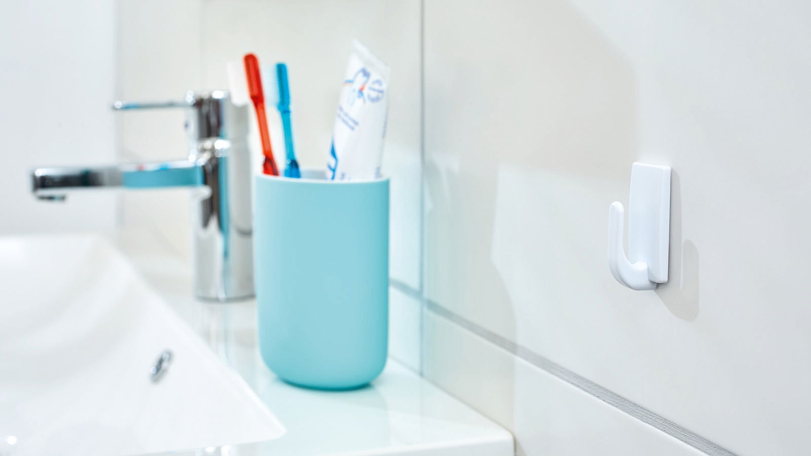 Badezimmeransicht mit einem weißen tesa Powerstrips Haken Small an der Wand, neben einem Waschbecken und einem Becher mit Zahnbürsten.