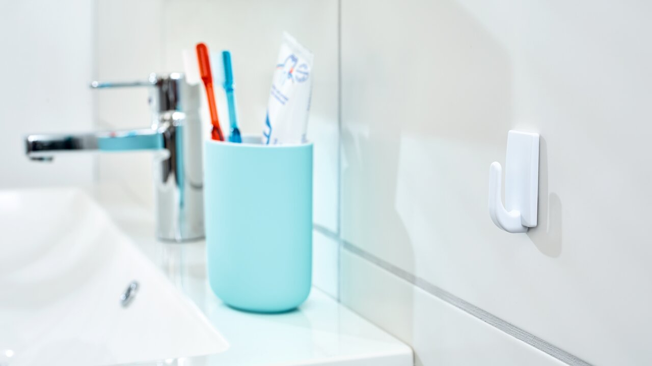 Badezimmeransicht mit einem weißen tesa Powerstrips Haken Small an der Wand, neben einem Waschbecken und einem Becher mit Zahnbürsten.